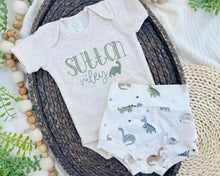Load image into Gallery viewer, An oatmeal beige bodysuit with the names 'Sutton Riley' embroidered on the front and a pair of blue shorts with a dinosaur pattern. Both items are laid out flat on a neutral background.