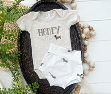 Load image into Gallery viewer, Baby onesie and shorts set with &#39;Henry&#39; and dachshund design on a wooden surface with decorative elements.
