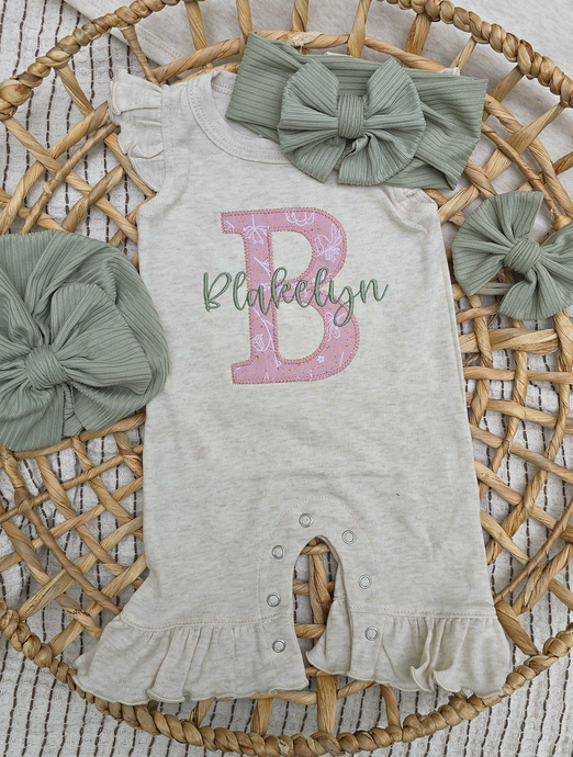 Gray romper with pink letter 'B' and name on a wicker tray with green bows.