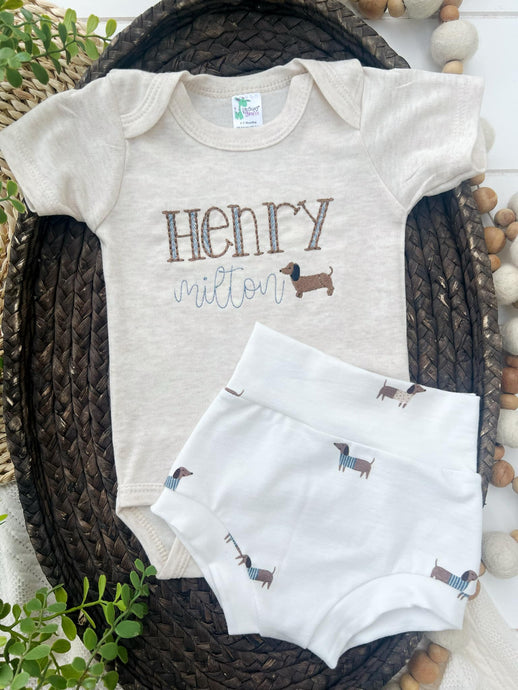 Baby onesie with 'HENRY milton' text and dachshund design, paired with a matching baby blanket on a woven basket.