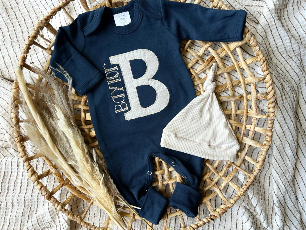 Navy blue baby onesie with a large white letter 'B' and matching hat on a woven basket.