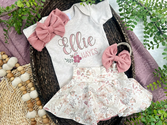 Baby outfit with floral dress, white onesie with 'Ellie Hayes' text, and pink headbands on a woven basket with greenery.