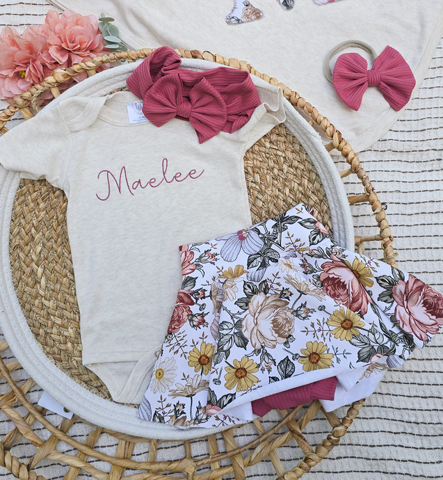 Baby outfit with floral design, name 'Maelee', and accessories on a woven mat.